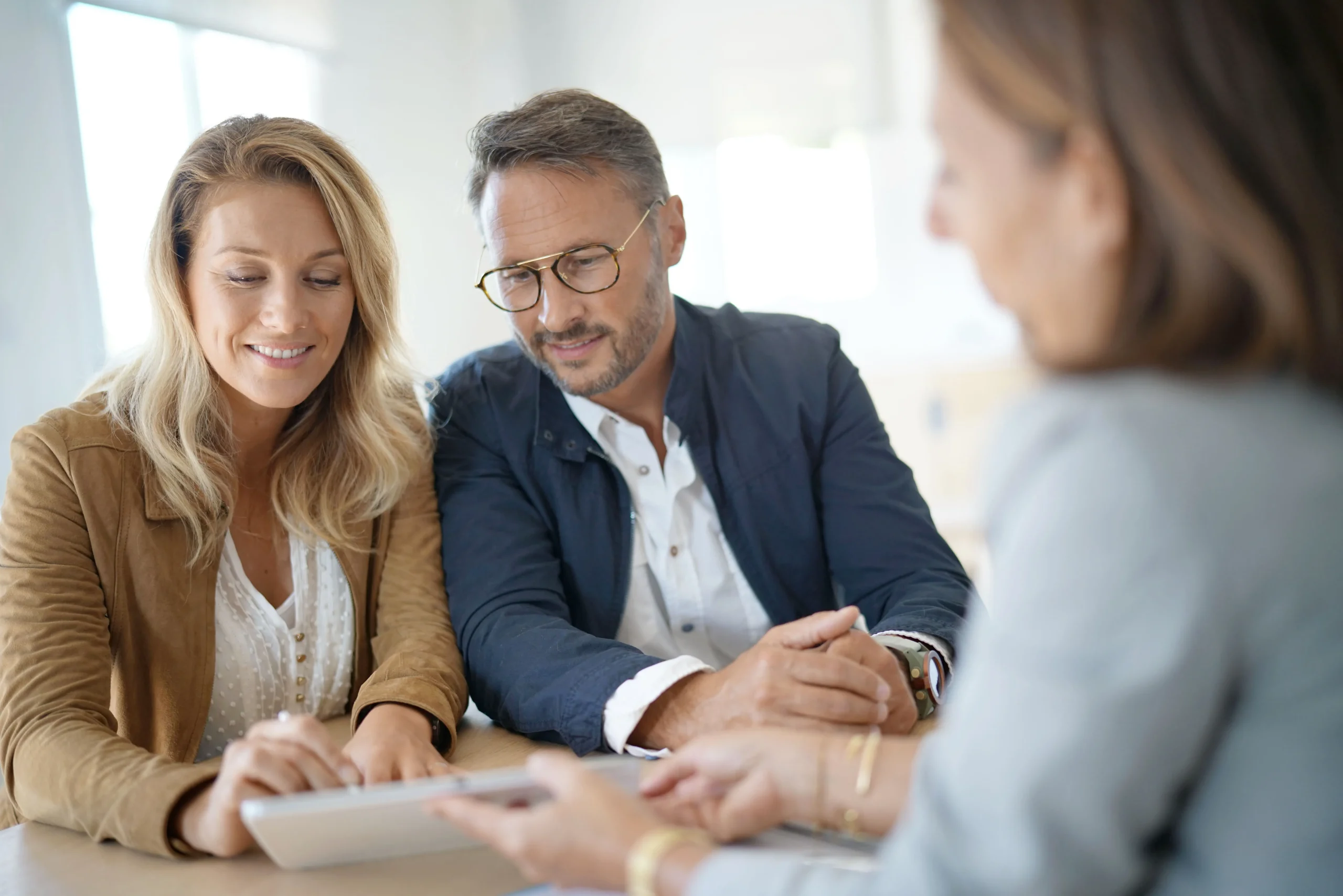 A couple sits at a table reviewing a tablet with a professional, while the advisor gestures toward the screen, suggesting a financial or consultation meeting.