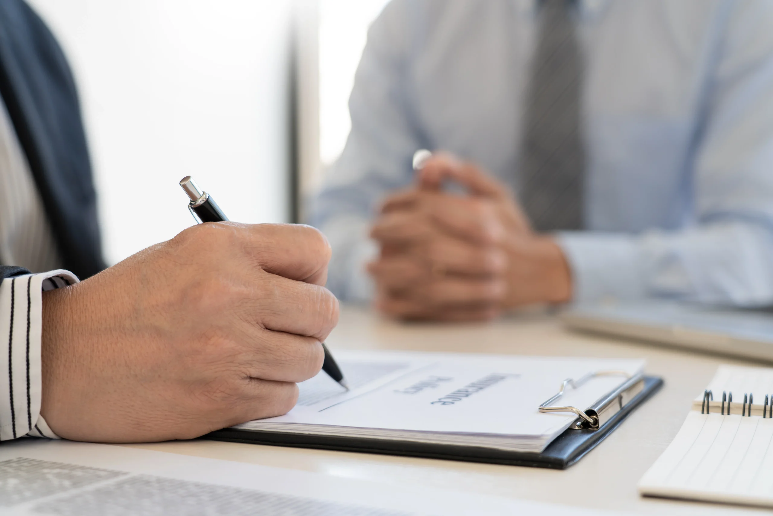 A person signs a document on a clipboard while another person sits across the table with hands clasped, suggesting a formal meeting or agreement.